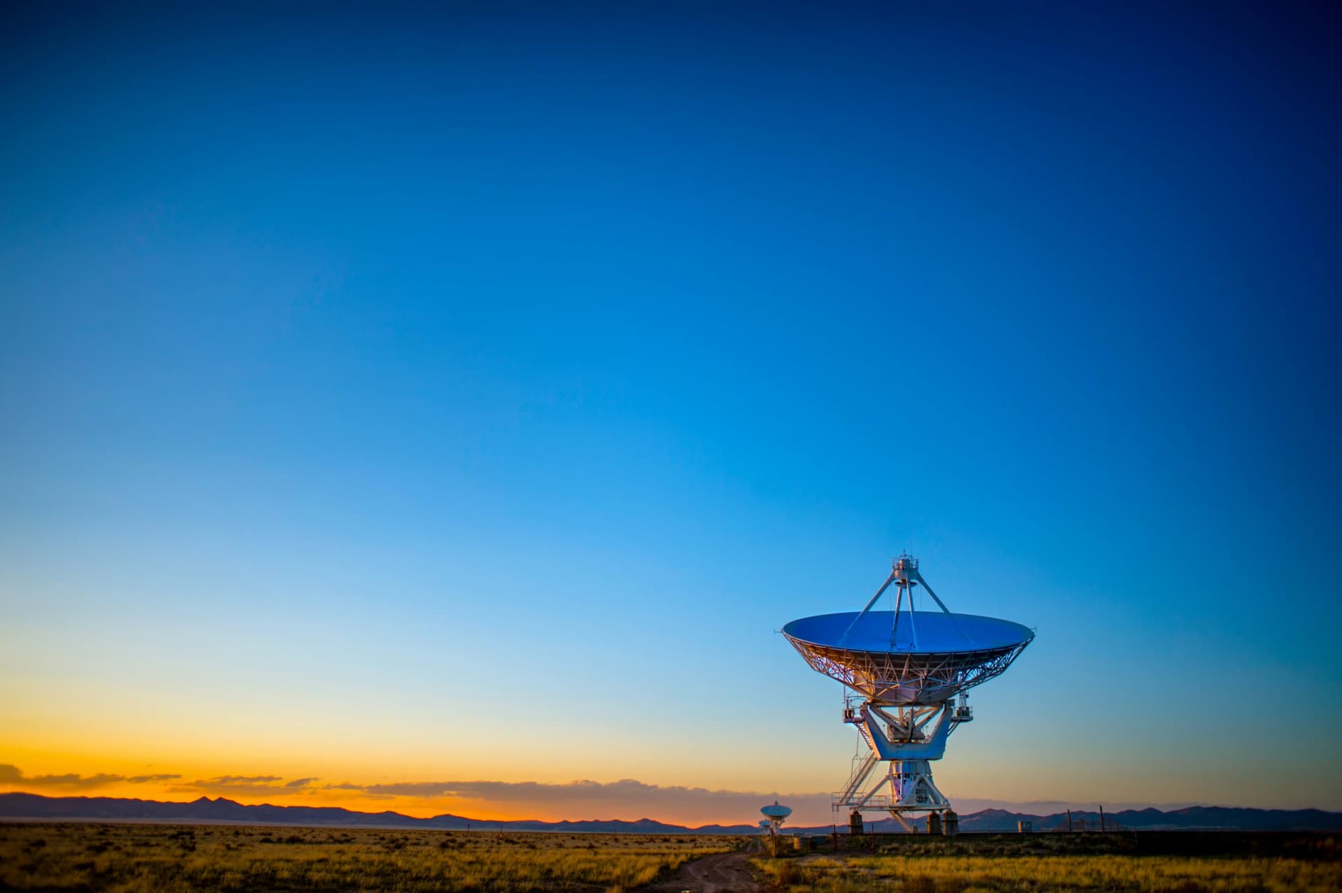 Large radio telescope dish standing on open grassland under a deep blue sky with an orange sunset glow on the horizon