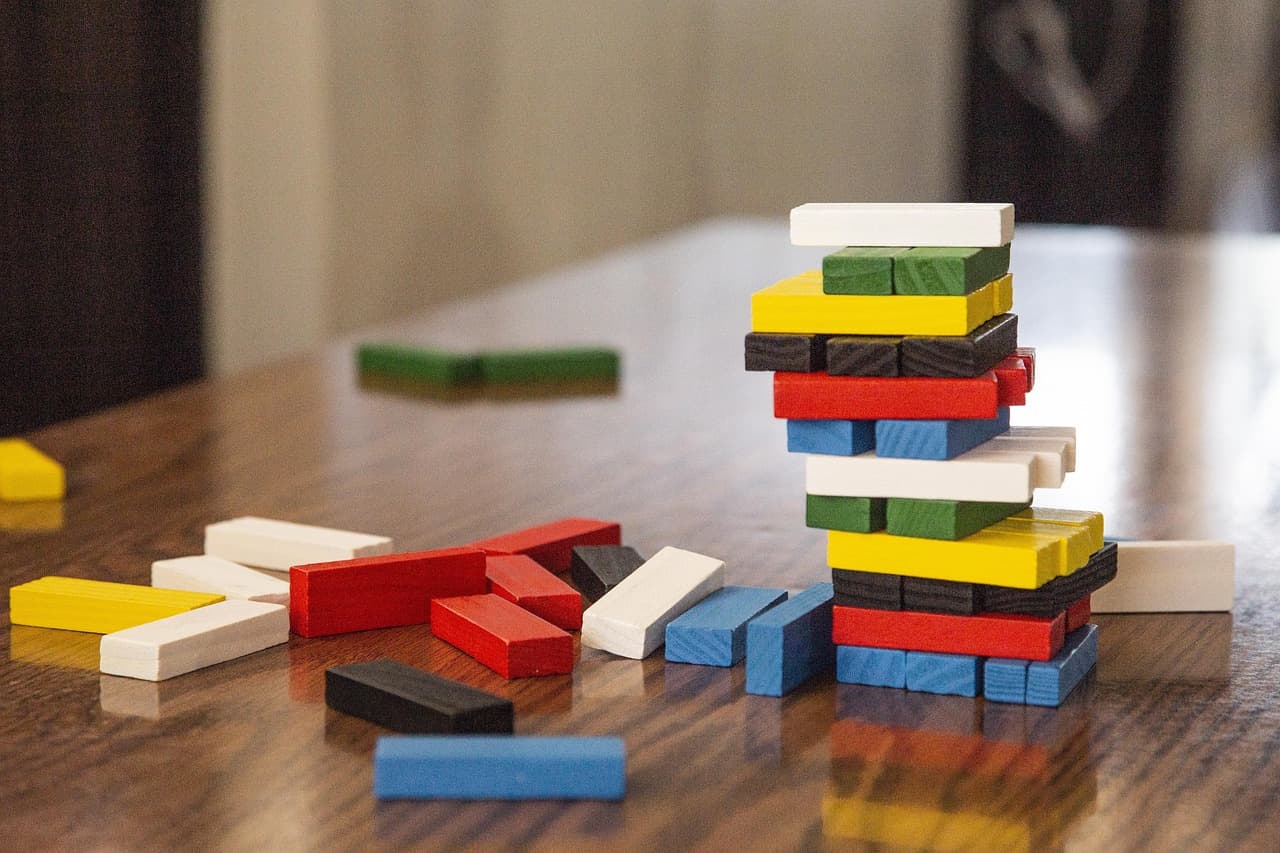 A partly toppled tower of colorful wooden Jenga-style blocks on a polished table, with loose red, blue, white, yellow, and black blocks scattered around it