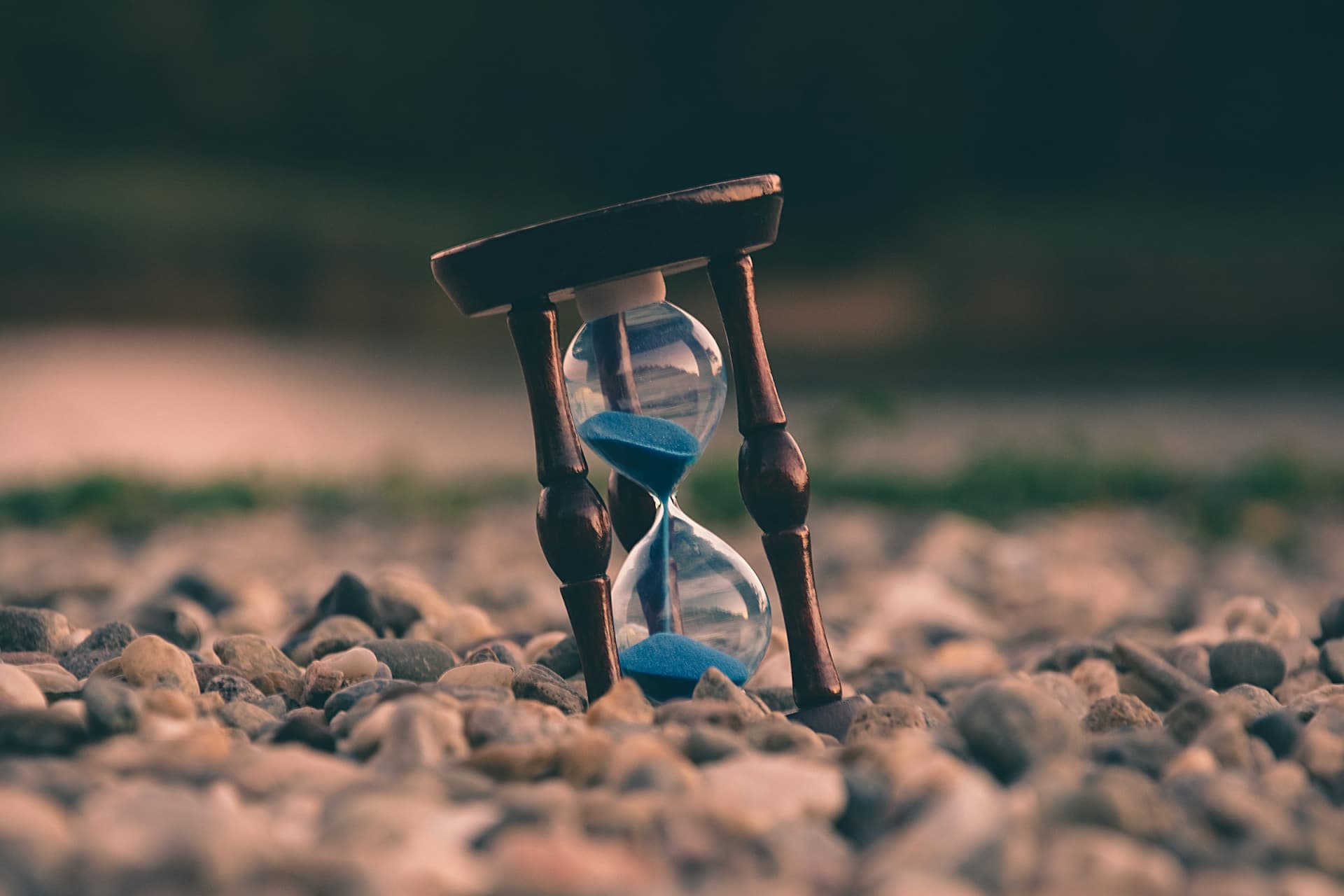 A wooden-framed hourglass with blue sand running out, standing among pebbles on the ground